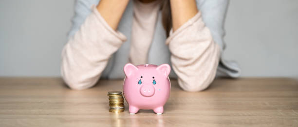 Sad girl leaned on the table, covering her face with hands, in the foreground a crying piggy bank with painted tears. Woman lost her savings, was fired, and is experiencing financial difficulties.