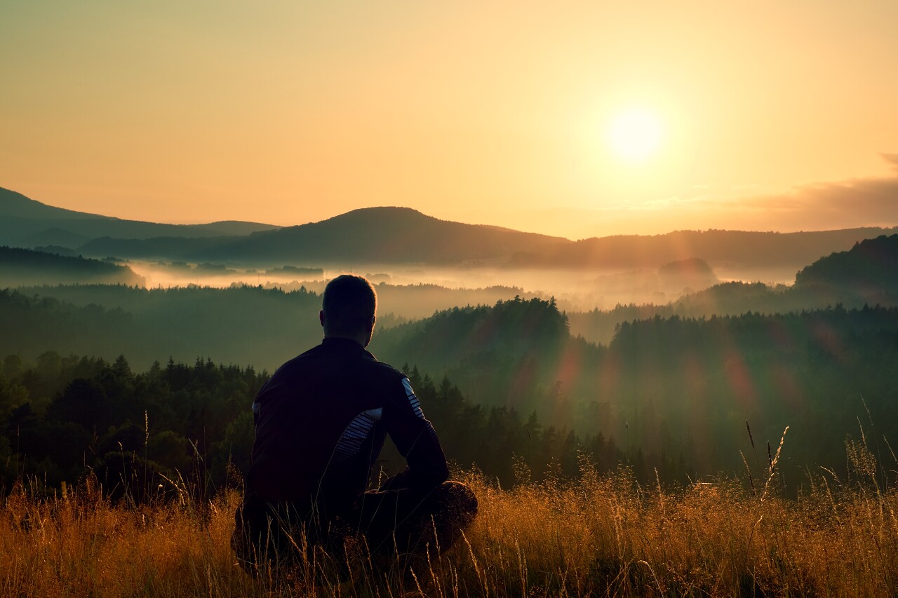 Hiker in squatting position in high grass meadow enjoy the colorful sunrise scenery